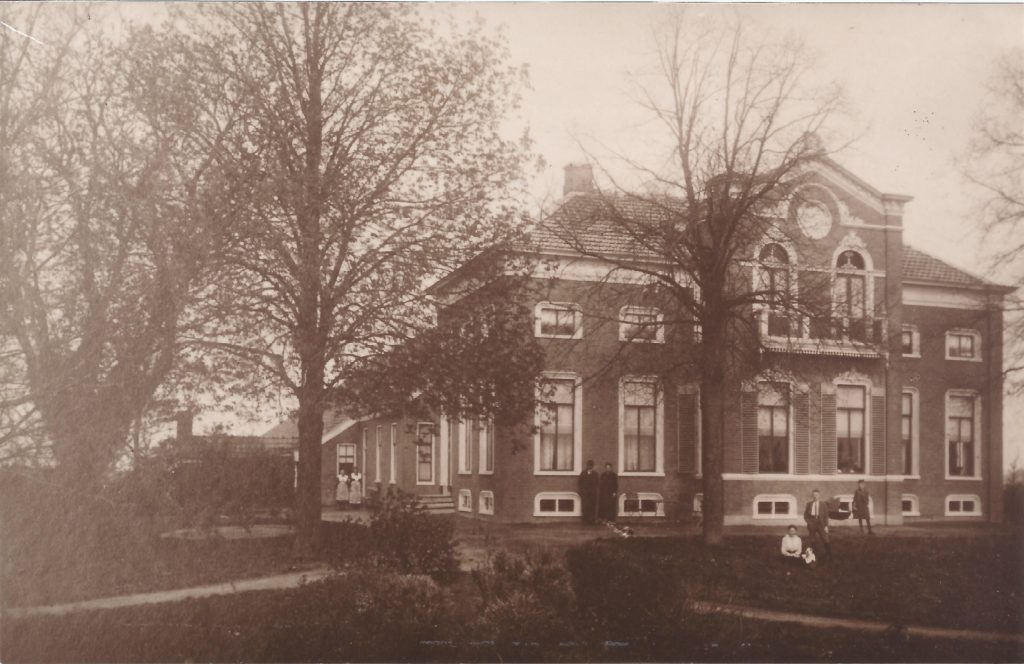 Large two story house, with a family in the foreground, and servants to the back. Old black and white photo , sepia toned. 