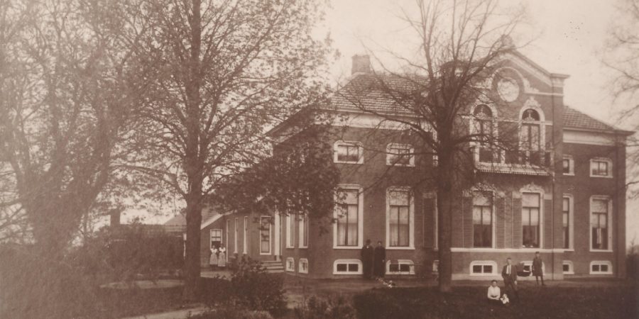 Sepia toned old photograph featuring a large two story house. The family is in the foreground, servants are to the back.